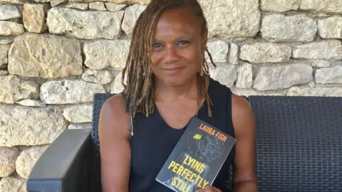 Michael Wild Laura Fish sits on a grey wicker garden chair, smiling and holding a book entitled in yellow lettering Lying Perfectly Still and with her name in orange lettering as author. She is wearing a dark vest top and has her brown hair in lots of thin braids. 