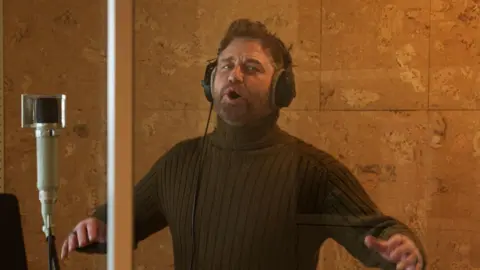 Sky Cinema Gerard Butler stands in a sound booth wearing a khaki turtle neck. His mouth is slightly open looking like he is singing and his hands raised up. 