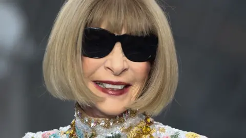 Getty Images A close up headshot of Anna Wintour smiling, wearing sunglasses. She has a bob haircut and is wearing a high collar embroidered dress studded with crystals.