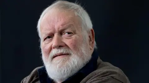 Getty Images Michael Longley has short white hair and beard and is wearing a brown cardigan over a dark blue shirt.