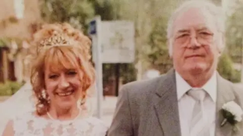 Family photograph Karen Teasdale-Robson in a white dress and veil smiling next to her father Brian Robson, an older man with white hair and glasses wearing a suit, tie and a carnation button hole.