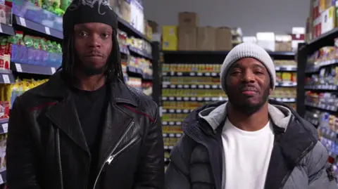BBC Krept and Konan are seen standing side-by-side facing the camera in an aisle of their supermarket.
