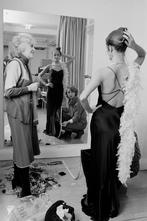 jean fennell at a costume fitting, wearing a black backless dress and feather boa looking in the mirror