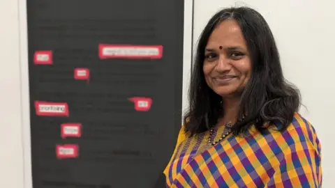 Abhi Arumbakkam A woman with dark hair and wearing a blue and yellow checked shawl, standing in front of a framed body of text. The text is too blurred to read, but there are seven words throughout that are highlighted and outlined in red.