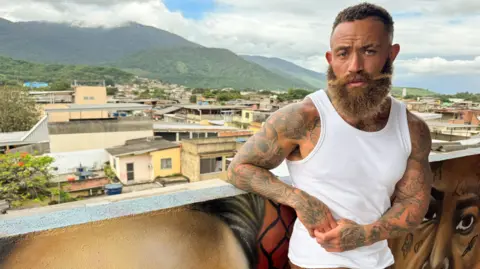 BBC/True North A man with a beard wearing a white top stands on a balcony in a favela in  Brazil