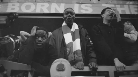 Leon Omondi A black and white photo taken from below shows Gabin, Tony,  and other fans. Tony is wearing a Chelsea scarf and looking directly at the camera. Other fans, including Gabin, are watching the game with stressed and shocked looks on their faces.