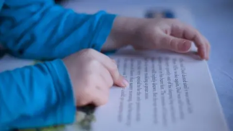 Getty Images A close-up photograph of a child reading a book. The child's hands and the book are visible and the child is using a finger to follow the words on the page.