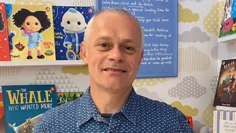 Handout Duncan Cooper has short grey hair. He is wearing a blue and white patterned shirt with a black T-shirt underneath. He is stood in his bookshop with children's books on white shelves behind him.