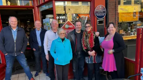 Script Haven A group of men and women stand outside a bookshop, which is painted red. They all smile at the camera.