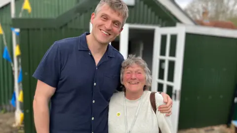 Jon Wright/BBC Director Callum Berridge and writer Mary Onions stand together in front of a small, green, corrugated metal building. There are blue and yellow flags behind them.