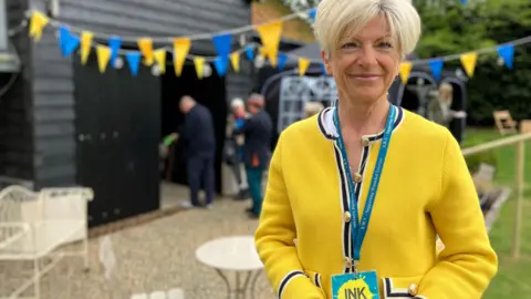 Jon Wright/BBC Julia Sowerbutts, in a bright yellow top, stands with an INK lanyard around her neck. there are blue and yellow flags in the background and a queue of people entering a barn.