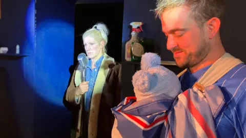 Jon Wright/BBC A woman stands wearing a sheepskin coat and speaking into a microphone, closer to the camera a man has a baby in a sling. He is wearing an Ipswich Town football shirt and the baby is draped in an Ipswich flag.