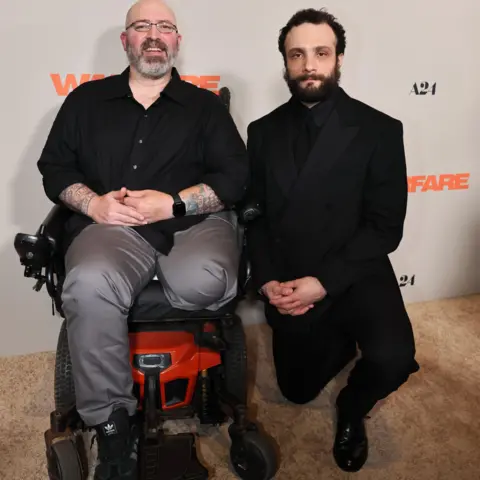Getty Images Veteran Elliott Miller in smart black and grey clothes in a wheelchair - one of his legs has been amputated - and Cosmo Jarvis next to him in smart, black clothes