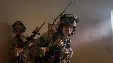 Murray Close Will Poulter and D'Pharaoh Woon-A-Tai in army uniforms holding guns on set