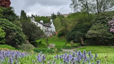 Cumbria PR Rydal Mount, a large white country cottage with a grey roof and several chimney stacks, stands in the background of the image, surrounded by trees and grass. A number of bluebells can be seen in the foreground.