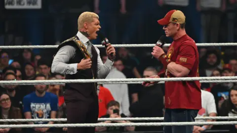 Getty Images Cody Rhodes and John Cena, two men, holding and speaking into microphones during a WWE promo in the ring. Cody is wearing a grey shirt, black waistcoat and trousers with a WWE title over his shoulder, while Cena is wearing a red tshirt, blue denim shorts and and a red and light brown hat.