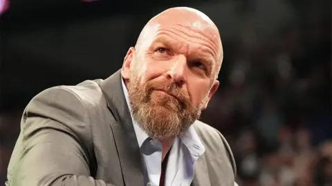 Getty Images Triple H, wearing an open-collared light blue shirt and grey blazer, looking out at the crowd, leaning over the wrestling ring ropes.