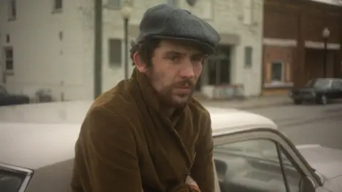 Mastermind Movie Inc Josh O'Connor in 1970s flat cap and brown coat in a still from The Mastermind