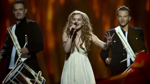 Getty Images emmelie de forest in a white dress sings with a man playing a bass drum on either side of her