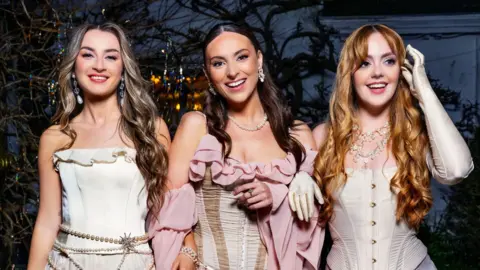 the trio remember monday in front of a tree. the woman of the left is blonde, the woman in the middle brunette and the woman on the right is a redhead. they are wearing white corsets, and the woman in the middle is also wearing pink ruffles