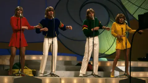 bucks fizz performing at the 1981 eurovision song contest. the band stands in a row, with the two women on the outside. the woman on the length is wearing a red thigh-high dress, and the woman on the right a yellow one. the men are wearing white trousers, with the one of the left in a blue jumper and the one on the right in green. the women's discarded long skirts lie on the ground behind them
