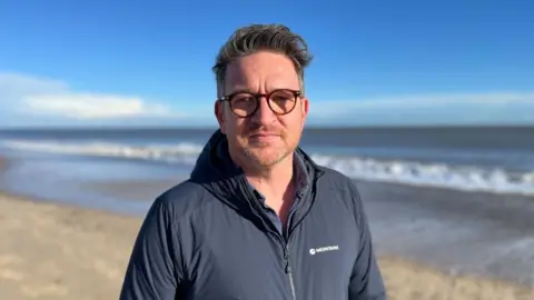 Shaun Whitmore/BBC Prof Ben Garrod, with round tortoise-shell glasses, and medium length dark hair swept over to his left, stands by the sea in Great Yarmouth. He has a stubbly beard and moustache, and he is wearing a blue jacket. The sky behind him is blue with a couple of clouds at low level.