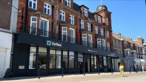 Andrew Turner/BBC The Place, Great Yarmouth. It is a four storey building with dormers and a domed tower with a flagstaff atop. The upper storeys feature white painted windows and bay windows on the first floor. The logo of The Place, and other signage adorns the fascia. A woman in yellow is walking past, and other shops are visible in the distance.