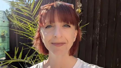 Katrin Oldridge Katrin Oldridge has red coloured hair, and is smiling, looking directly at the camera. She is wearing a white top, to the left of her head is a wind chime, and she is standing near a brown boarded fence in a garden. Behind her is a bamboo-type plant and a plastic greenhouse.