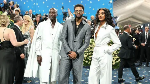Getty Images Ozwald Boateng stands on the left of a trio, with his two children, in a white suit and cape at the Met Gala. In the centre is his son, in a grey suit, and on the right, in white, is his daughter. Celebrities can be seen mingling in the background.