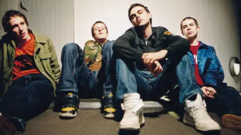 Bernice King Hard-Fi, shot from a low angle, sit on a plain carpeted floor, wearing early-2000s combat gear-style fashion