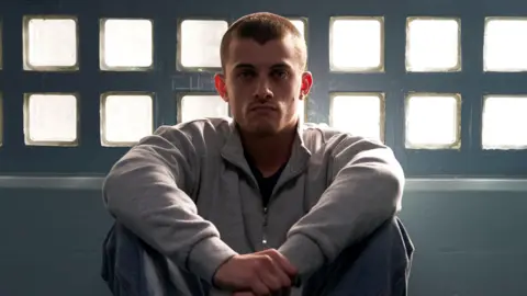 BBC/Hera/Enda Bowe Jake Dunn plays Liam - he is pictured here sitting down, wearing a light grey top and dark trousers.