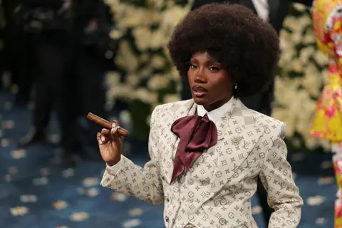 Getty Images Doechii wears a custom smoking jacket, crimson cravatte and a cigar, while posing on the red carpet of the 2025 Met Gala