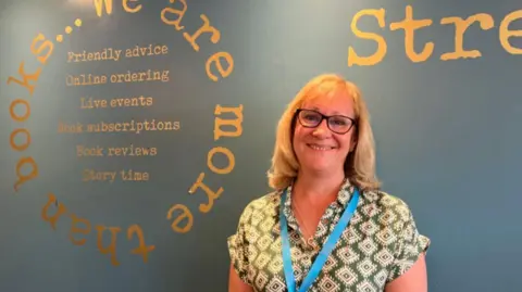 BBC Dawn Brewer, who owns Word on the Street bookshop in Ashstead, Surrey. She is blonde and is wearing glasses and a green and white patterned shirt. 