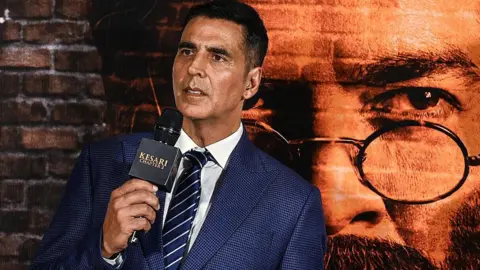 Getty Images Akshay Kumar seen wearing a blue suit with a poster of his film Kesari Chapter 2 in the background