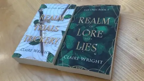 Two books side by side on a wooden table. One is white with a sword with the title 'Realm, Trials and Trickery. The other features green leaves and the title ' Realm of Lore and Lies'. 
