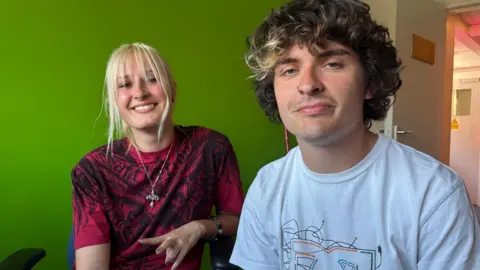 BBC A young woman with blond hair and young man with brown, curly hair sit in a room with bright green walls. The young woman wears a burgundy top with a intricate black pattern inspired by heavy metal artwork and a silver pendant on a long chain.
