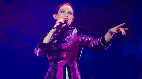 Getty Images Sophie Ellis-Bextor is performing in stage wearing a reflective purple coat.