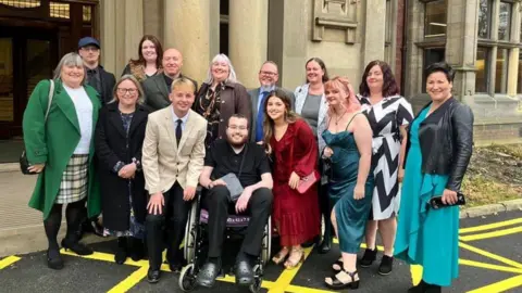 Supplied Mr Swales in the middle of family photograph on his graduation day in November 2024. He is smiling and wearing black trousers and a black t-shirt. His brother-in-law, Stephen Jefferson, is next to him in a beige suit jacket, white shirt, red tie and black trousers. He is smiling and has blond hair. Everyone else is also dressed formally.