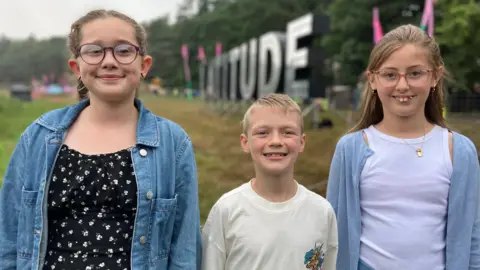 Jon Wright/BBC Two girls and a boy stand in front of a festival sign that reads Latitude