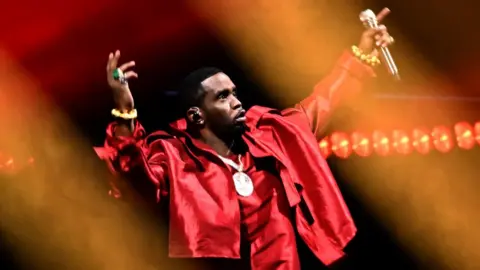 Getty Images Combs on stage wearing a bright red shirt and jacket and a large gold medallion with both of his arms gesturing in the air during the 2023 MTV Video Music Awards.