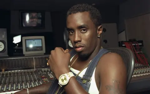 Getty Images Sean Combs wearing denim dungarees, a white vest and a gold watch, sitting at a mixing desk in a recording studio in 1997