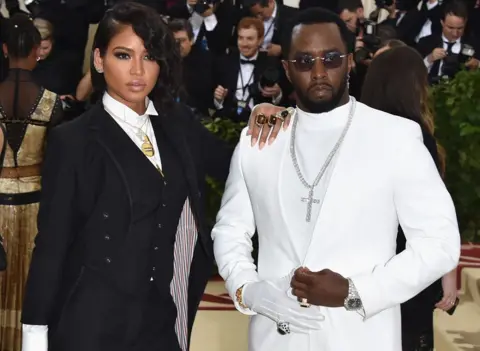 Getty Images Cassie Ventura and Sean Combs posing for photographers at the 2018 Met Gala. She's wearing a black suit and he's wering an all-white suit with one white glove and a large jewelled cross on a chain around his neck