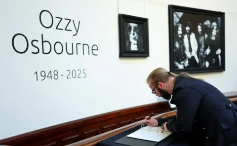 Reuters A man with a dark suit, ponytail and beard signing a book of condolence in front of a wall with large wording saying "Ozzy Osbourne 1948-2025"