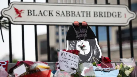 EPA A 2D face of Ozzy with a Black Sabbath hat, sunglasses, a bat necklace and a cross necklace placed there by fans, in front of a sign saying Black Sabbath Bridge