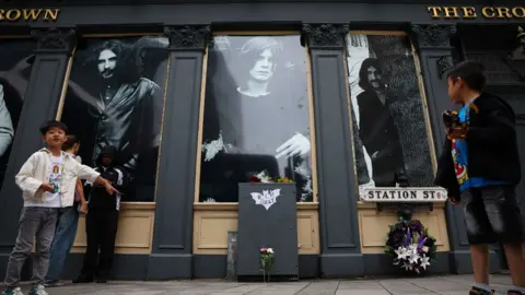 EPA Exterior of the Crown pub with black and white photos of different band members in each window, with a few tributes on the front and a few people standing in front of it