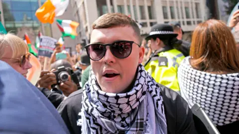 PA Media A man wearing glasses and a keffiyeh, stands outside court - in the background dozens of supporters, some waving Irish flags, can be seen