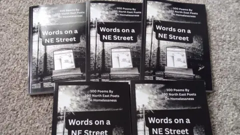 Andrew Mears Five of the anthologies are laid out on a grey carpet. The front cover is a black and white picture of a street with a wooden bench on it, along with a lam post and a window to a building where light shines through. The title of the book is: "Words on a NE Street", "100 poems by 100 North East Poets on Homelessness".