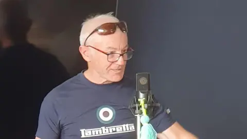 Maureen Waters Andrew Mears is standing in front of a microphone and speaking animatedly. He has short grey hair and is wearing black glasses. He also has a pair of sunglasses resting on top of his head.