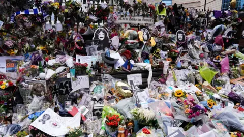 PA Media A huge pile of bunches of flowers, messages and other items left in tribute on a bench. Behind the bench are four cut-outs featuring each face of the Black Sabbath members. Behind them is a sign that says "Black Sabbath Bridge".