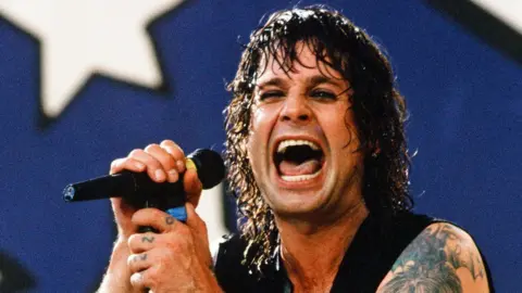 Getty Images Ozzy Osbourne - with long black, wet hair and a sleeveless black top - has his mouth wide open as he sings into a microphone, during his younger days.